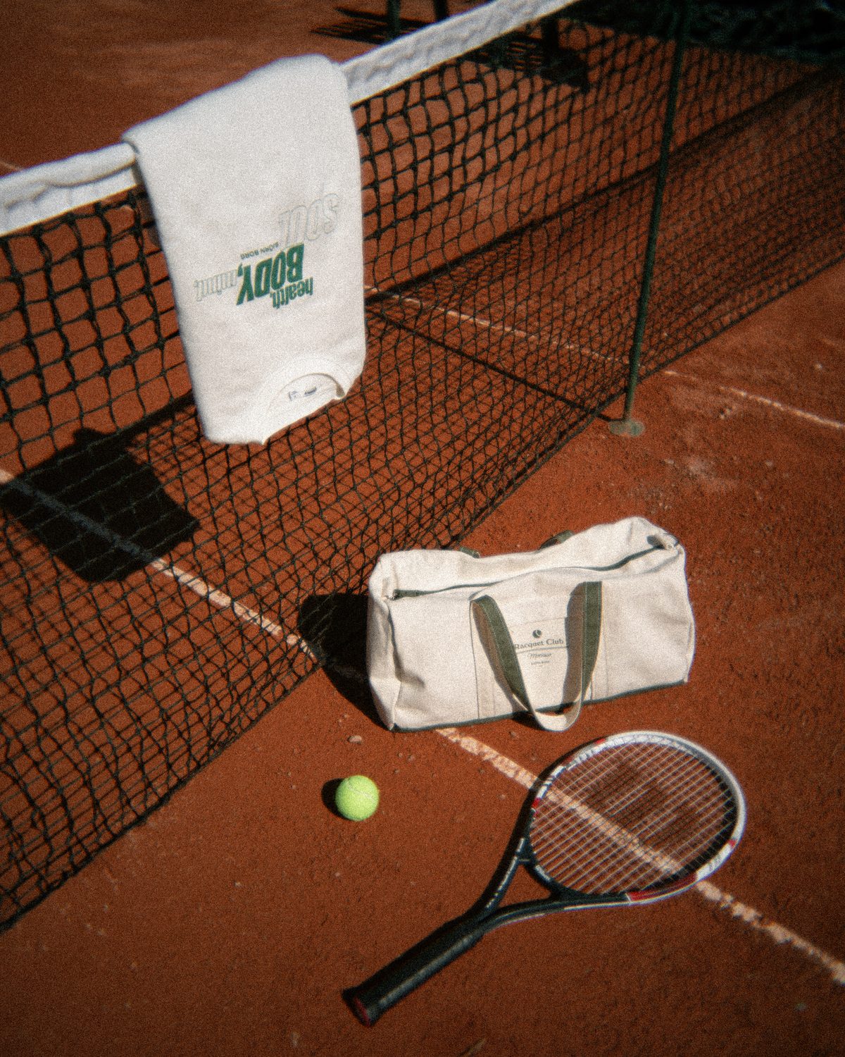 A styled frame on a clay tennis court: a towel on the net, a bag, a ball and a racket