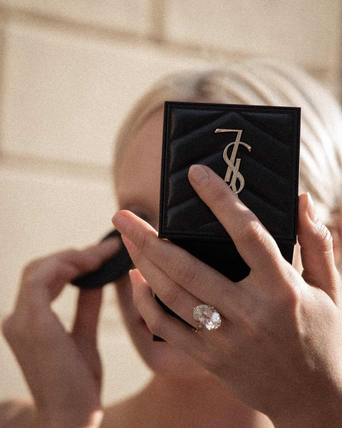 A hand with a large ring holding a quilted compact mirror up to the light
